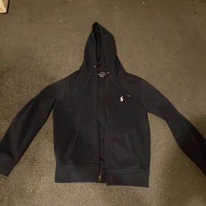 Marinblå huvtröja från Polo Ralph Lauren - Säljer en stilren marinblå huvtröja från Polo Ralph Lauren med dragkedja och det klassiska logotypbroderiet på bröstet. Den är i nyskick och har använts 1-2 gånger. Tröjan har långa ärmar och en praktisk huva med dragsko. Perfekt för en avslappnad look eller till vardags.