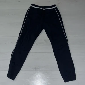 Svarta mjukisbyxor från Hugo Boss - Snygga svarta mjukisbyxor från Hugo Boss med vita detaljer längs sidorna. Byxorna har en bekväm midwaist och snörning i midjan för perfekt passform. Perfekta för en avslappnad stil. Väldigt bekväma!!!