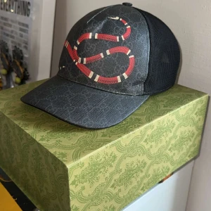Svart Gucci keps med röd orm - Säljer en snygg svart keps från Gucci med ett coolt rött ormtryck. Kepsen har ett diskret mönster och justerbar rem baktill. Perfekt för att ge din outfit en lyxig touch! Storlek M