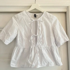 Vit blus från H&M - Säljer en söt vit blus från H&M med knytband framtill och korta ärmar. Blusen har en luftig passform och är perfekt för varmare dagar.