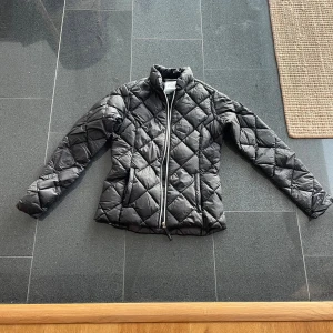 Svart quiltad dunjacka från McKinley - Säljer en svart quiltad dunjacka från McKinley. Jackan har en dragkedja framtill och är perfekt för kyligare väder. Den är designad med långa ärmar och har en stilren look. Obetydligt använd, inga fläckar eller märken, nyskick.