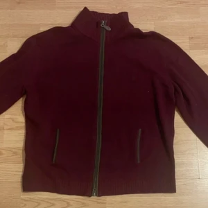 Vinröd tröja med dragkedja från Genfins - Säljer en vinröd tröja från Genfins med dragkedja framtill. Tröjan har en hög krage och är tillverkad i en ullblandning för extra värme. Perfekt för kyligare dagar.