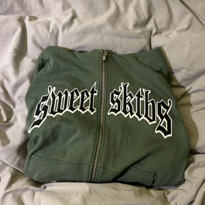 Grön Zip hoodie från Sweet SKTBS - Säljer en grön hoodie från Sweet SKTBS med dragkedja och stor logga på framsidan. Den har en bekväm passform med långa ärmar och en praktisk huva med snörning. Perfekt för en avslappnad stil. Tröjan är i storlek L och har en relaxed passform. Köpt för 700kr för ca 6 månader sedan och sparsamt använd under kortare period