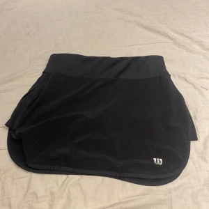 Svart kjol från Wilson - Snygg svart kjol från Wilson, perfekt för tennis eller annan sport. Kjolen har en stilren design med en liten logga i nederkant. Tillverkad i ett bekvämt material som ger bra rörelsefrihet.