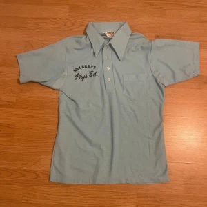 Ljusblå pikétröja  - Säljer en ljusblå pikétröja från Florida med texten 'Hillcrest Phys. Ed.' broderad på bröstet. T-shirten har en klassisk krage och knappar framtill. Materialet är en blandning av bomull och polyester, vilket gör den både bekväm och hållbar.