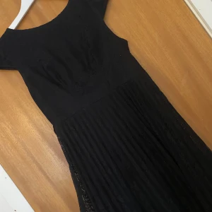 Svart klänning med spetsdetaljer - Elegant svart klänning med spetsdetaljer och rund hals. Perfekt för en stilren look. Klänningen har en vacker struktur och är ärmlös, vilket ger en sofistikerad känsla.