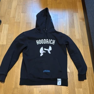 Svart hoodie från Hoodrich - Säljer en svart hoodie från Hoodrich med tryck av vita fåglar och text på både fram- och baksidan. Hoodien har en stor ficka framtill och justerbar huva med dragsko. Perfekt för en avslappnad stil.