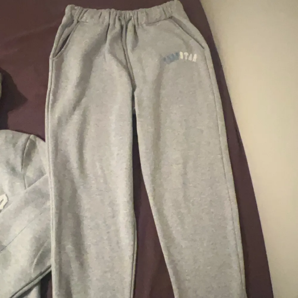 Säljer en grå hoodie från Trapstar med text i blått och vitt på bröstet. Den har en klassisk design med dragsko i huvan och ribbade muddar vid ärmslut och nederkant. Perfekt för en avslappnad stil.. Hupparit & Collegepaidat.