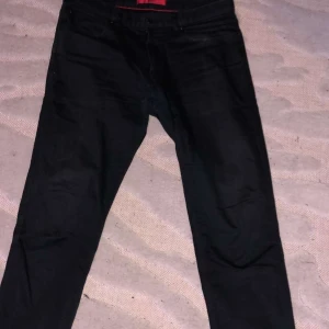 Svarta byxor från Hugo - Snygga svarta byxor från Hugo med en klassisk design. Byxorna har en röd etikett på insidan och baksidan, vilket ger en stilren kontrast. Perfekta för en stilren look. 