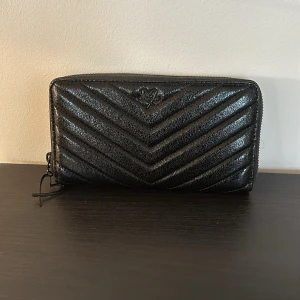 Svart Victoras secret plånbok - Snygg svart plånbok med quiltat mönster och dragkedja. Den har flera fack för kort och sedlar, samt en hjärtformad dragkedja. Perfekt för att hålla ordning på dina tillhörigheter med stil.