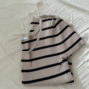 Beige och svart randig ribbad topp - Säljer en snygg beige topp med svarta ränder. Den är ribbad och har korta ärmar, perfekt för en stilren look. Toppen har en rund halsringning och är i ett mjukt material.