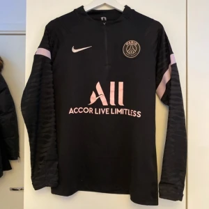 PSG tröja - Säljer en svart långärmad tröja från Nike med Paris Saint-Germain-logga på bröstet. Tröjan har en dragkedja vid halsen och rosa detaljer samt texten 'Accor Live Limitless' framtill. Perfekt för fotbollsfans!