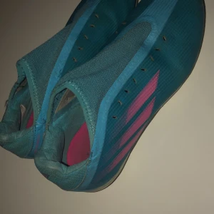 Blå och rosa fotbollsskor från Adidas - Snygga blå fotbollsskor från Adidas med rosa detaljer. Skorna har en strömlinjeformad design utan snörning för en tight passform. Perfekta för snabba rörelser på planen.