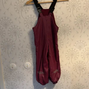 Vinröda hängselbyxor för barn - Säljer ett par vinröda hängselbyxor för barn med justerbara svarta hängslen. Byxorna har broderade detaljer vid bensluten och är perfekta för vinterlek.