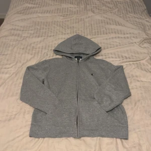 Grå hoodie från Ralph Lauren - Säljer en klassisk grå hoodie från Ralph Lauren med dragkedja och huva. Perfekt för en avslappnad stil. Den har långa ärmar och en liten logotyp på bröstet. Passar bra till både jeans och joggers.
