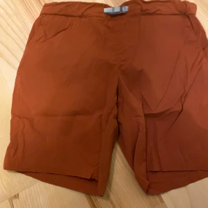 Orange shorts från Houdini - Säljer ett par orange shorts från Houdini i damstorlek M. De har en praktisk justerbar rem i midjan för perfekt passform. Perfekta för sommarens äventyr!