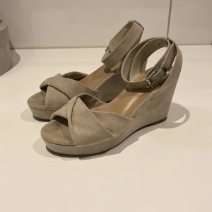 Beige kilklackssandaler - Snygga beige sandaler med kilklack och korsade remmar framtill. De har en justerbar ankelrem med spänne för perfekt passform. Perfekta för sommarens alla tillfällen. Klackhöjd är 8cm.