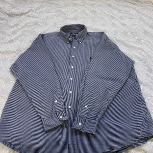 Randig skjorta från Ralph Lauren - Snygg randig skjorta från Ralph Lauren i blått och vitt. Skjortan är långärmad och har klassiska knappar framtill. Tillverkad i 100% bomull för en bekväm passform. Perfekt för en stilren look.
