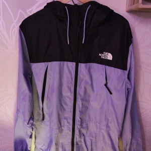 Svart och ljusblå vindjacka från The North Face - Säljer en stilren svart och ljuslila vindjacka från The North Face. Jackan har en praktisk huva och dragkedja framtill. Perfekt för blåsiga dagar och har märkets logga på bröstet och ärmen.