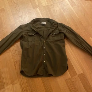Olivgrön overshirt från Barbour - Snygg olivgrön overshirt från Barbour i regular fit. Skjortan har långa ärmar och knappar framtill samt två bröstfickor med knappstängning. Perfekt för lager-på-lager-stil under svalare dagar.