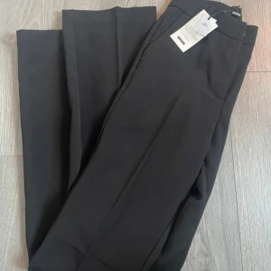 Svarta kostymbyxor - Snygga svarta kostymbyxor med hög midja och rak passform. De är helt nya och oanvända. Perfekta för en stilren look. Byxorna har en klassisk design med dragkedja och knapp framtill.