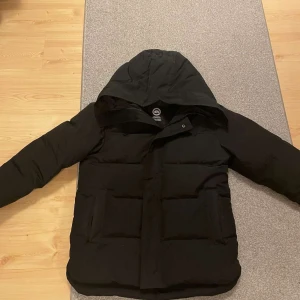 Svart dunjacka från Canada Goose - Säljer en svart dunjacka från Canada Goose med huva och dragkedja. Jackan har ett broderat märke på ärmen och är perfekt för kalla dagar. Den är långärmad och har en klassisk design som håller dig varm och bekväm.