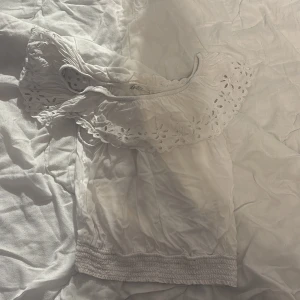 Vit blus med spetsdetaljer - Säljer en söt vit blus med vackra spetsdetaljer runt halsen. Blusen har korta ärmar och en elastisk nederkant som ger en fin passform. Perfekt för varma dagar!