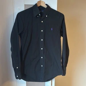 Svart skjorta från Ralph Lauren - Snygg svart skjorta från Ralph Lauren med klassisk krage och knappar framtill. Den har en broderad logga i lila på bröstet och långa ärmar med knappar vid ärmsluten. Perfekt för en stilren look.
