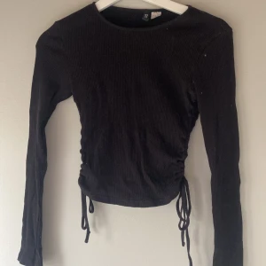 Svart ribbad tröja med knyt - Snygg svart ribbad tröja med långa ärmar och knyt i sidorna för en justerbar passform. Perfekt för en stilren look.