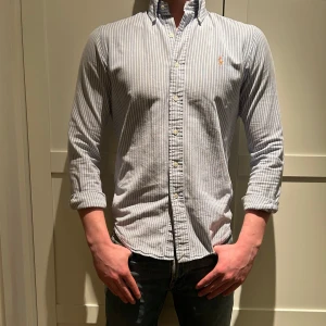 Randig skjorta från Polo Ralph Lauren - Snygg och stilren skjorta från Ralph Lauren i blå och vit randigt mönster. Skjortan har långa ärmar och en klassisk krage. Perfekt för en dressad look med en liten broderad logga på bröstet.