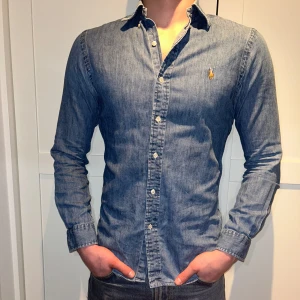 Blå jeansskjorta från Ralph Lauren - Snygg blå jeansskjorta från Ralph Lauren med klassisk krage och knappar framtill. Skjortan har en broderad logotyp på bröstet och långa ärmar. Perfekt för en stilren look.