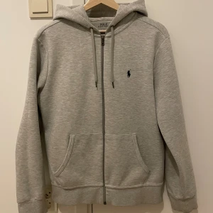 Grå hoodie från Polo Ralph Lauren - Säljer en stilren grå hoodie från Polo Ralph Lauren med dragkedja och justerbar huva. Tröjan har en broderad logotyp på bröstet och praktiska fickor framtill. Perfekt för en avslappnad look. Inga skador förutom 2 små fläckar på fickan som knappt synns.