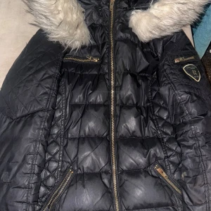 Svart quiltad jacka från Rockandblue - Snygg svart quiltad jacka från Rockandblue med pälskantad huva och dragkedja. Jackan har flera praktiska fickor med dragkedjor och ett stilrent märke på ärmen. Perfekt för kyliga dagar. Lagad, se sista bild. Syns inte så tydligt