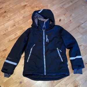 Svart vinterjacka Åre från Lager 157 - Säljer en svart vinterjacka från Lager 157 med grå detaljer. Jackan har en justerbar huva och flera praktiska fickor med dragkedjor. Den är utrustad med Hydratex-teknologi för extra skydd mot väder och vind. Perfekt för kalla dagar! ❄️