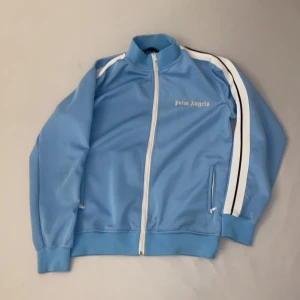 Blå track jacket från Palm Angels - Snygg ljusblå track jacket från Palm Angels med vita detaljer och dragkedja. Jackan har långa ärmar med vita ränder längs sidorna och en broderad logga på bröstet. Perfekt för en sportig look!