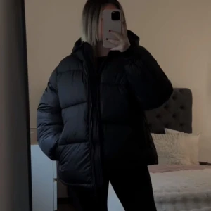 hm Svart trendig pufferjacka - Säljer en svart pufferjacka med huva. Jackan har en dragkedja framtill och är perfekt för kyliga dagar. Den är långärmad och har en bekväm passform. Storlek M, finns någon liten defekt samt att sminkfläck finns vid halsen men borde gå bort i tvätten. Hör av dig vid intresse❤️