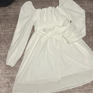 Vit klänning med puffärmar - Säljer en söt vit klänning med puffiga långa ärmar och en knytning i midjan. Klänningen har en romantisk stil med rynkade detaljer vid halsen och en luftig kjol. Perfekt för sommarens alla tillfällen!