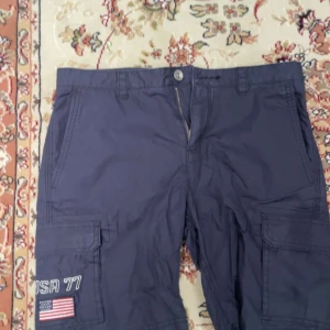Mörkblå shorts från Sail Racing - Snygga mörkblå shorts från Sail Racing med USA 77 och amerikansk flagga broderad på benet. Shortsen har flera praktiska fickor och en knappstängning fram. Perfekta för en avslappnad stil.