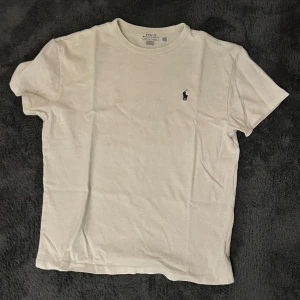 Vit t-shirt från Ralph Lauren - Säljer en klassisk vit t-shirt från Ralph Lauren med den ikoniska broderade loggan på bröstet. T-shirten är kortärmad och tillverkad i mjuk bomull. Perfekt för en stilren look.