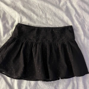 Svart kortkjol från Gina Tricot - Snygg svart kortkjol från Gina Tricot med små paljetter och volangdetaljer. Perfekt för att ge en stilren look med en twist. Passar bra till olika tillfällen och kan enkelt matchas med olika toppar. Säljer för att den är för li