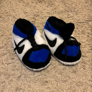 Sneakers tofflor - Mysiga tofflor som efterliknar Nike jordans i färgen blå. De har en fluffig yta i blått, svart och vitt med snörning framtill. Perfekta för att hålla fötterna varma och bekväma hemma!