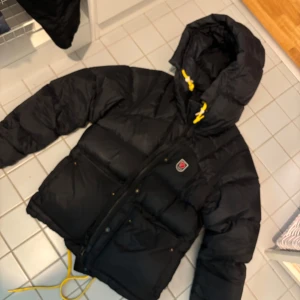 Svart dunjacka från Fjällräven - Säljer en svart dunjacka från Fjällräven, Expedition down lite Storlek S dammodell Inköpt på länna sport.Jackan har en huva med gula detaljer och dragkedja framtill. Perfekt för kalla dagar.Inköpt januari 2023 Nypris 8000