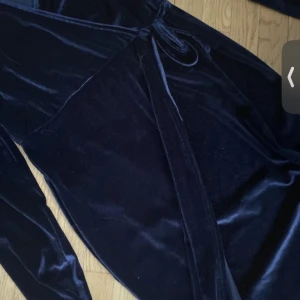 Mörkblå sammet klänning från Boohoo - Elegant mörkblå klänning i mjuk sammet från Boohoo. Klänningen har en omlott-design med knytning i midjan och långa ärmar. Perfekt för en stilfull look.