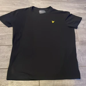 Svart t-shirt från Lyle & Scott - Snygg svart t-shirt från Lyle & Scott med en gul broderad logga på bröstet. Perfekt för en stilren och enkel look. Tillverkad i mjukt material för extra komfort.