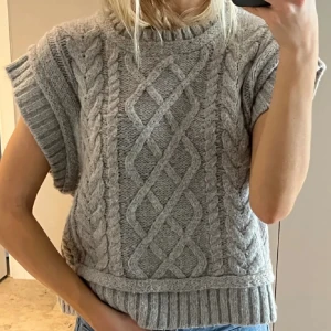 Grå stickad ärmlös tröja från neo noir - Säljer en snygg grå stickad tröja med flätmönster. Den är ärmlös och har ribbade kanter vid hals och nederkant. Perfekt för lager-på-lager-stil! (Prislappen sitter kvar helt oanvänd)
