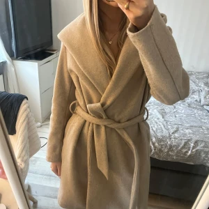 Beige kappa från Zara - Säljer en stilren beige kappa från Zara med knytbälte i midjan. Den har en elegant krage och långa ärmar, perfekt för kyligare dagar. Kappan är tillverkad i ett mjukt material som ger en lyxig känsla. Passar utmärkt för både höst och vår. Nopprig på vissa ställen men jag har en noppermaskin💕
