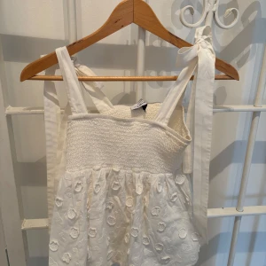 Vit klänning med blommönster - Charmig vit klänning med smockad överdel och axelband.