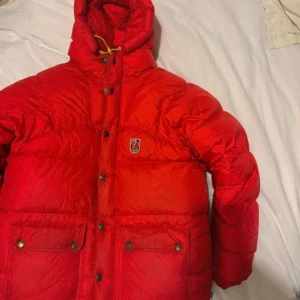 Röd dunjacka från Fjällräven - Säljer en snygg röd dunjacka från Fjällräven med huva och knappar. Jackan har en klassisk design med två stora fickor framtill och en broderad logotyp på bröstet. Perfekt för kalla dagar!