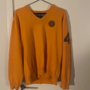 Orange tröja från GANT - Säljer en snygg orange tröja från GANT med v-ringning och ett tryck på bröstet samt siffran 4 på ärmen. Perfekt för en avslappnad stil med en touch av sportig elegans. Passar även M, Passar för en fin dag på golfbanan, kan byta också, 