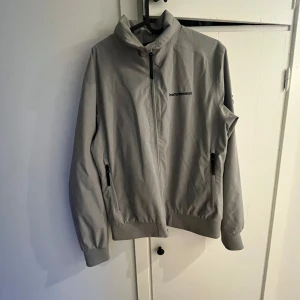 Grå jacka från Peak Performance - Säljer en stilren grå skal jacka från Peak Performance. Jackan har dragkedja och ribbade muddar vid ärmslut och nederkant. Perfekt för en sportig look.